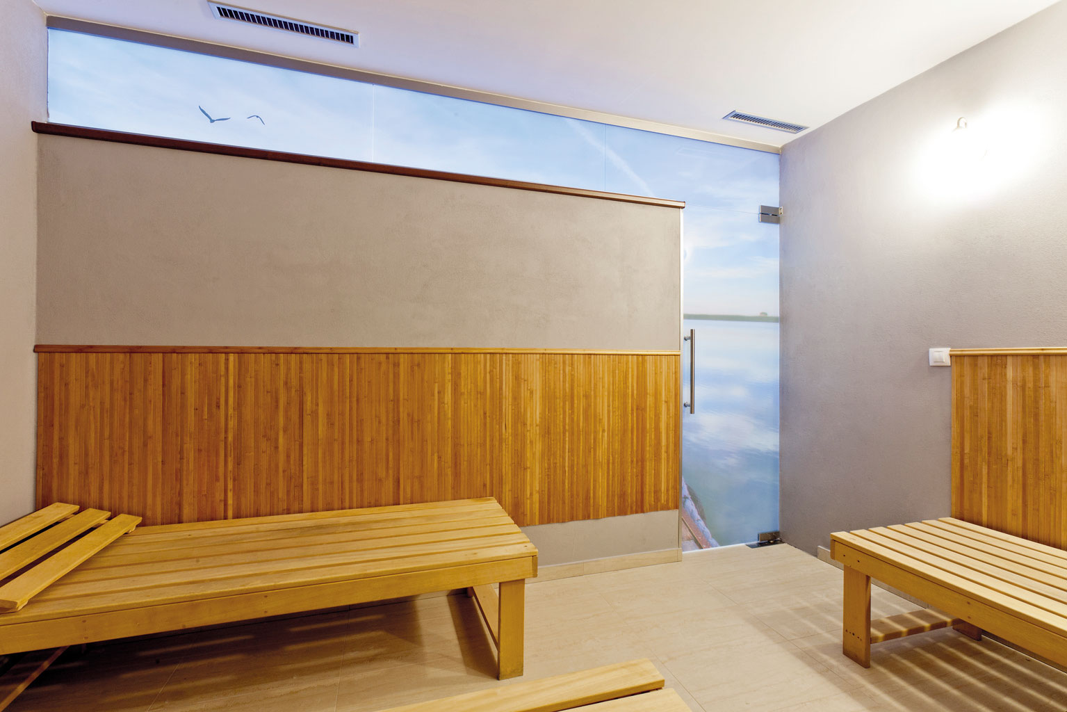 Pohľad z odpočivárne, kde celosklenené dvere a svetlík nechávajú presvitať dekor zapadajúceho slnka nad morskou hladinou zo susediacej miestnosti.